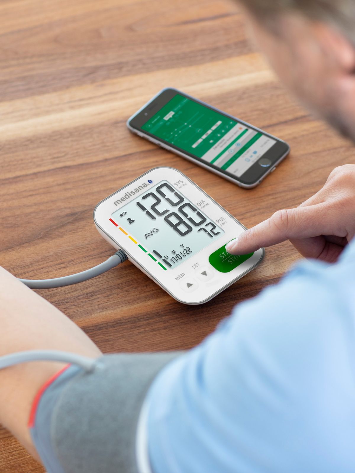 A person measures their blood pressure using a digital upper arm blood pressure monitor while a smartphone with a matching health app lies next to it.